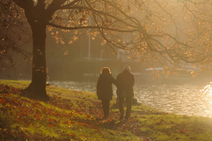 A couple walk arm in arm along a grassy riverbank under an autumn tree, with fallen red leaves and hazy golden light on the water