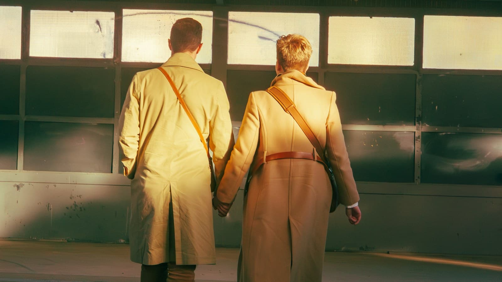 A couple in long camel coats, seen from behind, walk hand in hand through a passage lit by low golden sun