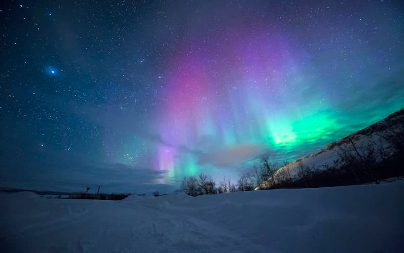 Grønt og lilla nordlys over et snødekt landskap med stjernehimmel.