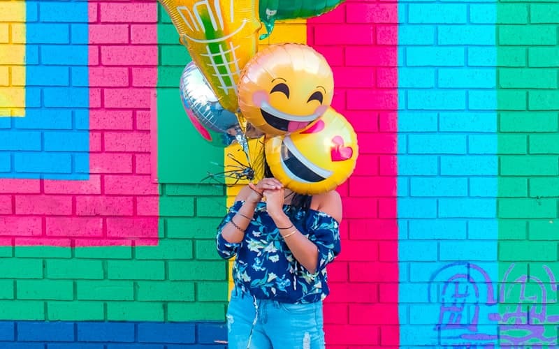 Someone in a blue floral top holds smiley balloons before a brightly painted brick wall.