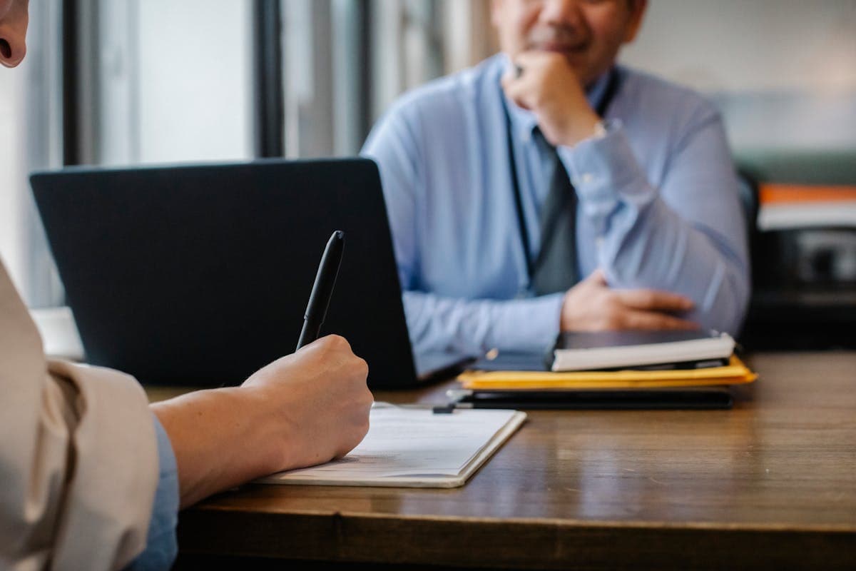 Someone writes on a document at a wooden table while a man in a blue shirt listens.