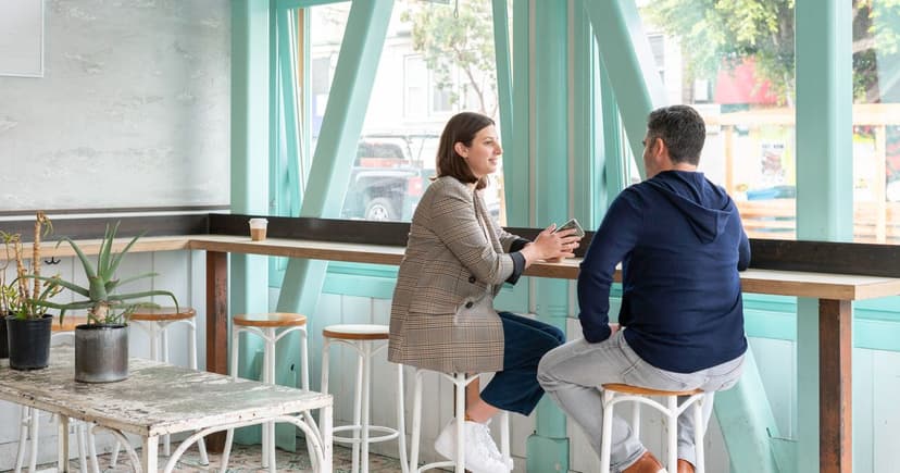 A woman in a checked blazer and a man in a navy hoodie talk over coffee at a bright cafe window counter.
