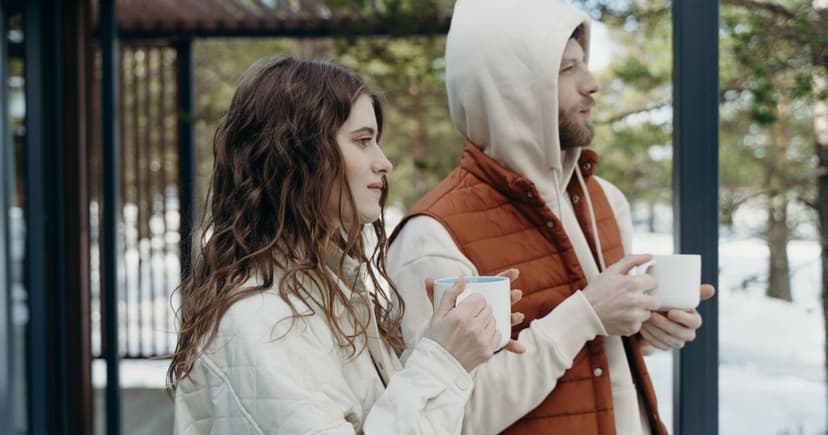 A woman and a man in quilted jackets hold white mugs on a covered deck, snow and pine trees behind.