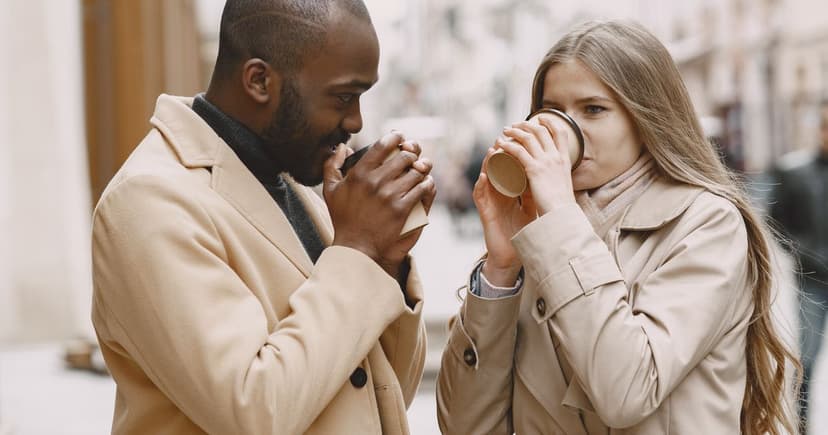 A man and a woman in camel overcoats stand close together outdoors, each sipping from a takeaway coffee cup on a cool day.