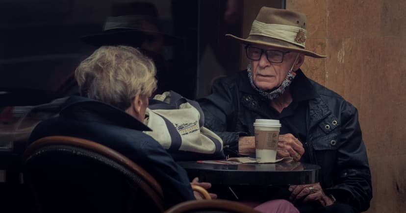 An older man in a wide-brimmed hat talks with a blonde woman over takeout coffee at a small sidewalk cafe table.