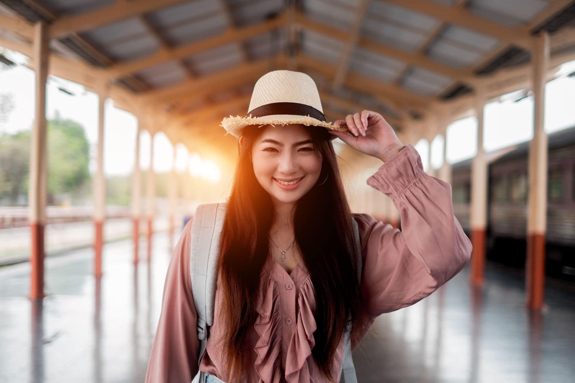 Eine lächelnde Frau mit Strohhut steht im warmen Licht auf einem Bahnsteig.