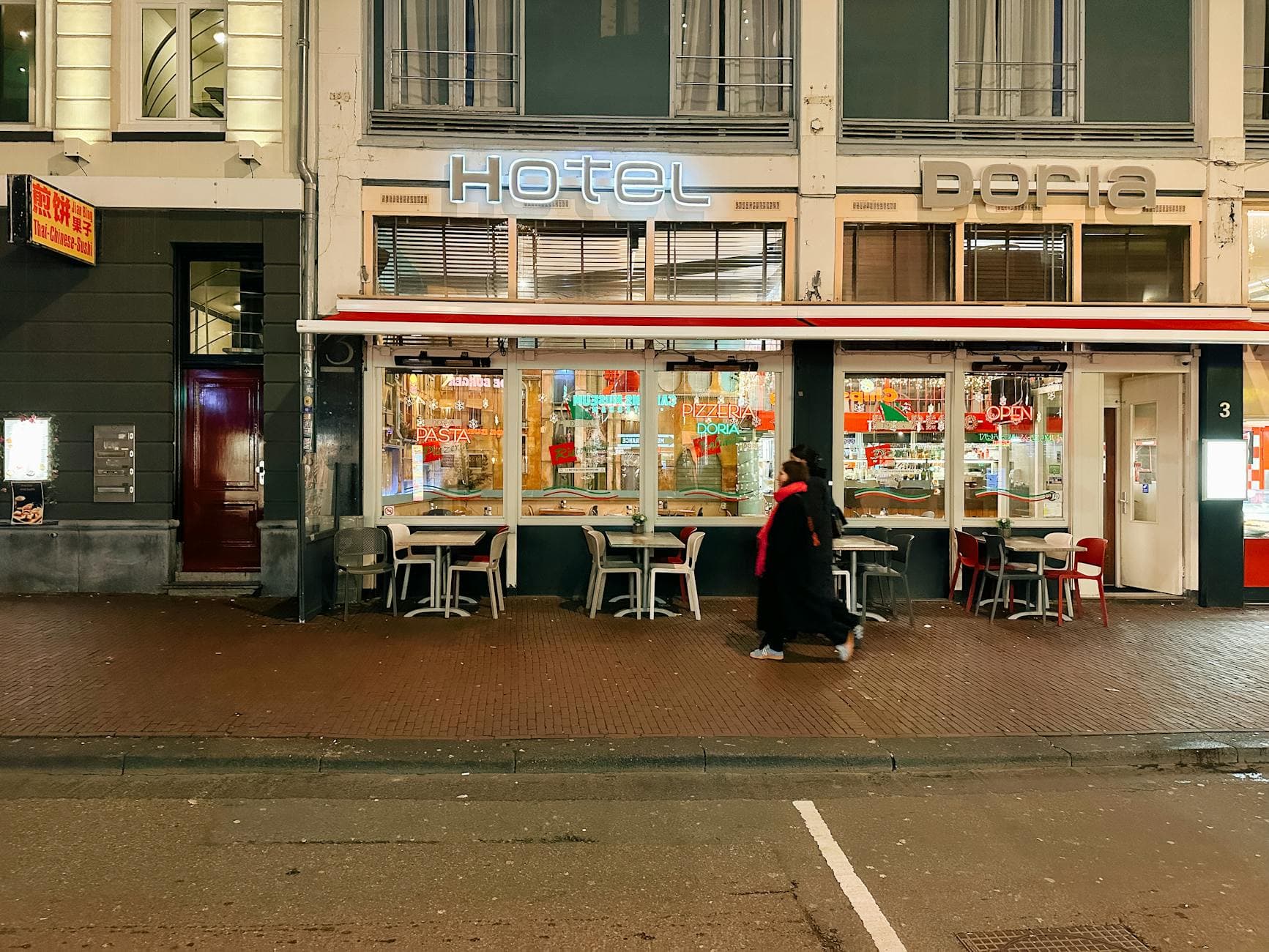 Iemand in zwarte jas met rode sjaal loopt 's avonds langs een verlichte pizzeria.