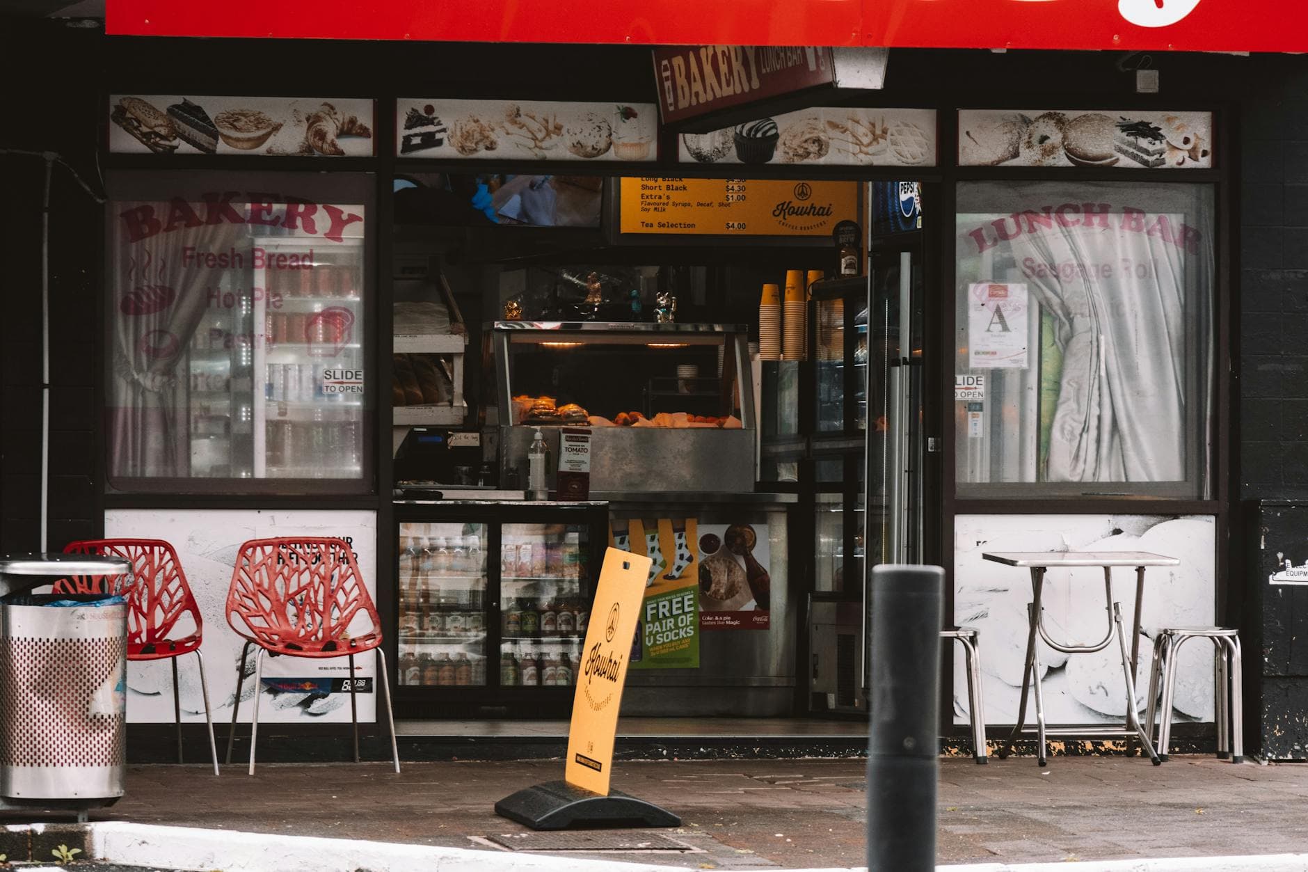 A bakery and lunch bar shopfront with red plastic chairs and a yellow sign on the footpath.