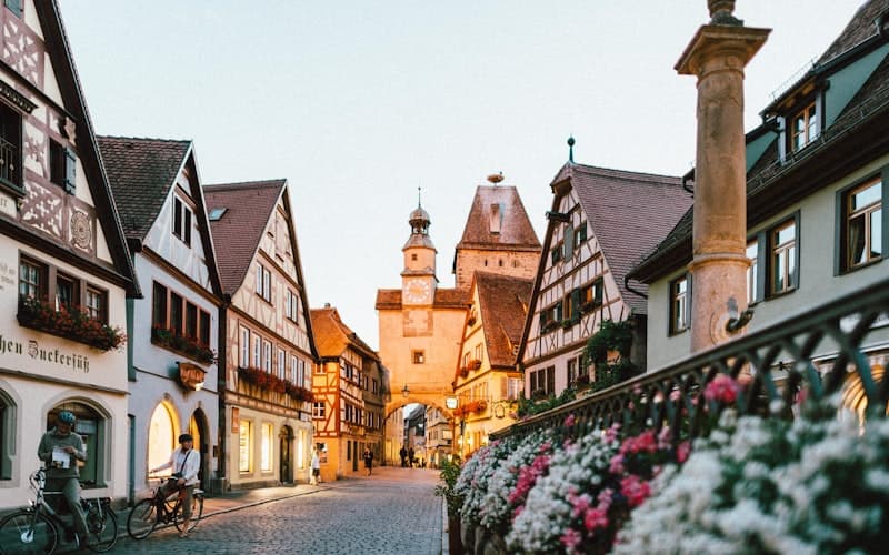 Fachwerkhäuser und ein Torturm säumen abends eine Gasse mit blühenden Blumen.