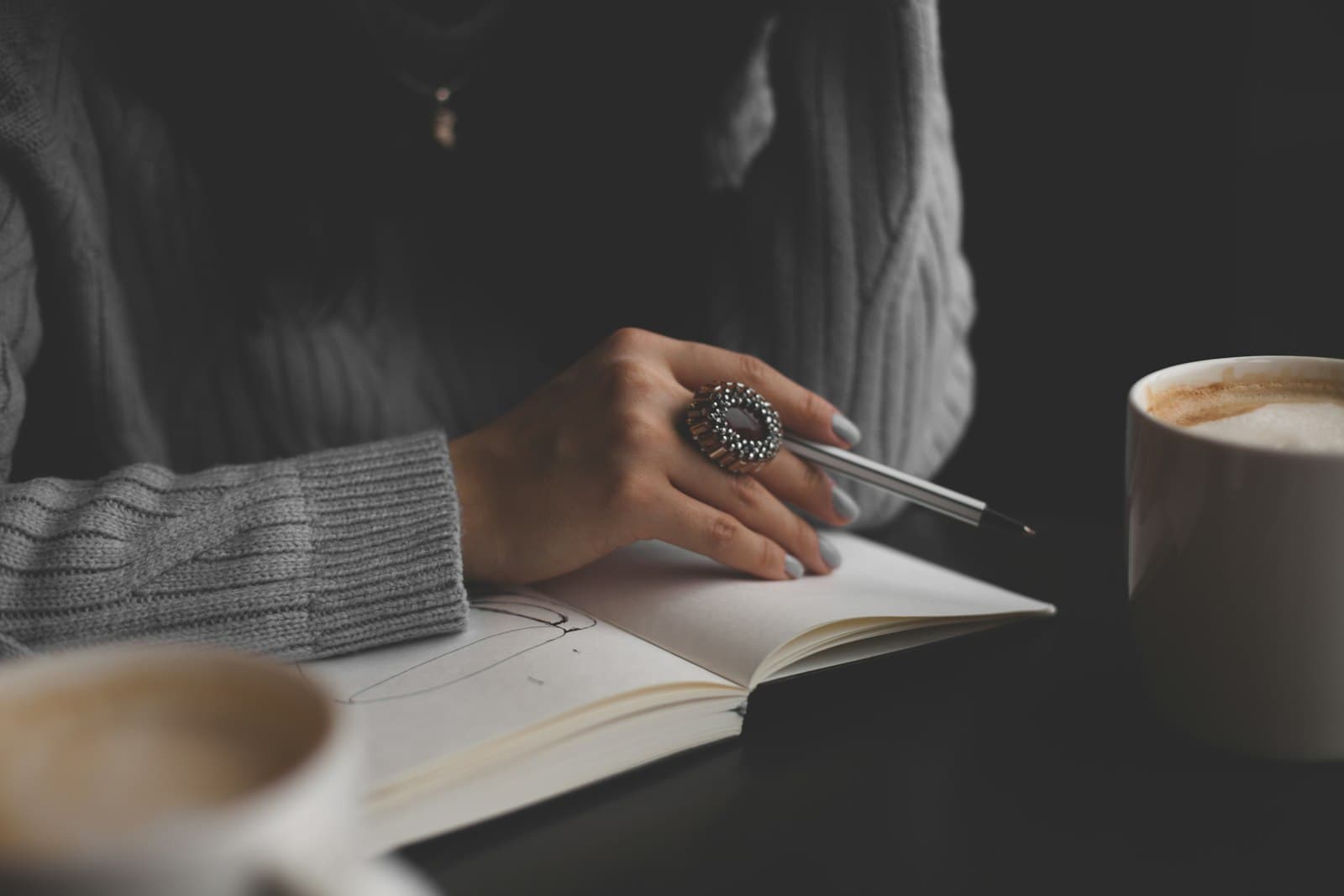 Hand mit großem Ring hält einen Stift über einem Notizbuch, daneben Cappuccino