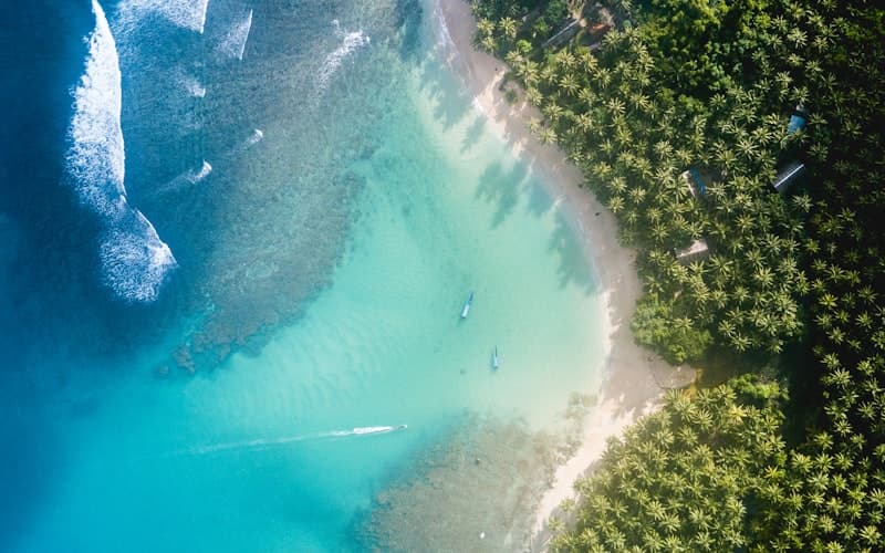 Flygbild över turkos lagun med vit sandstrand, palmer och små båtar i vattnet.