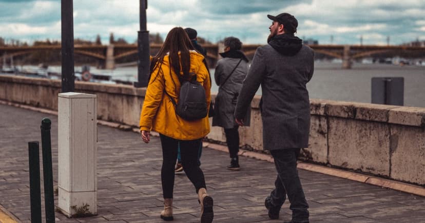 A couple in warm coats walk along a stone-walled waterfront promenade on an overcast day, with a bridge in the distance.