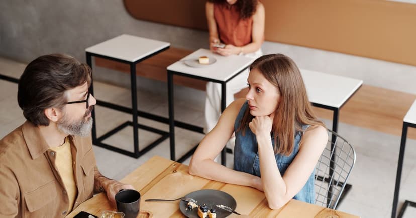 A grey-bearded man and a younger woman sit talking over coffee and dessert at a table in a bright, modern café.
