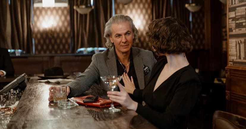 A silver-haired man with a whiskey tumbler talks across a wooden bar to a dark-haired woman holding a martini glass.