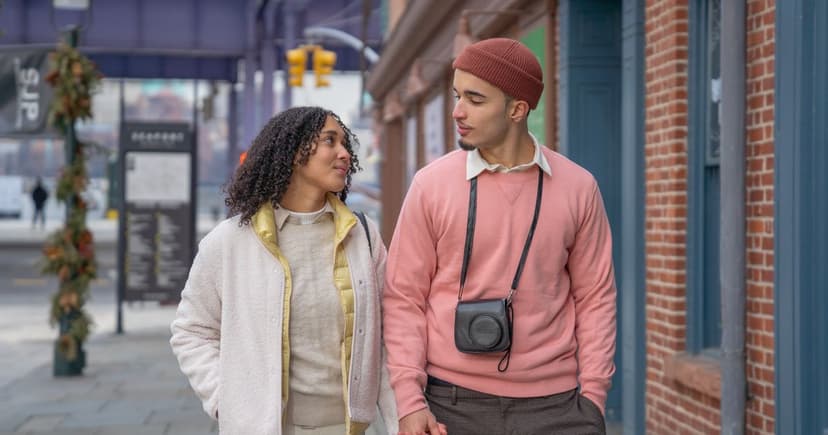 Un couple marche côte à côte sur un trottoir bordé de façades en brique rouge, en se regardant.