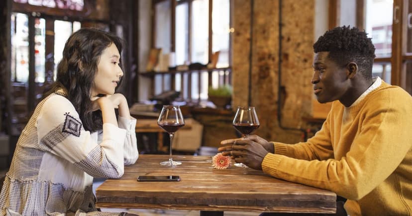 Un homme et une femme discutent face à face autour de deux verres de vin rouge, à une table en bois dans un bistrot aux murs de brique.