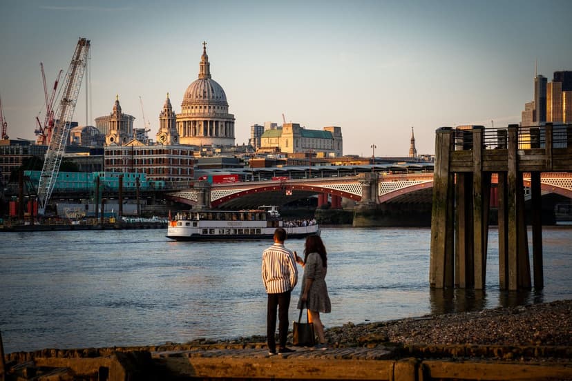 A couple stands together on the Thames foreshore at golden hour, watching a riverboat pass beneath Blackfriars Bridge with the dome of St Paul's Cathedral glowing behind them