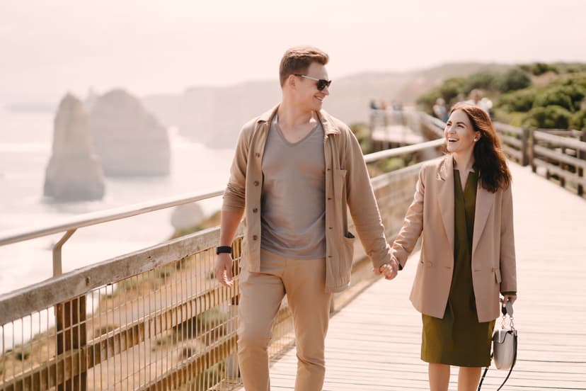 A laughing, stylishly dressed couple walks hand in hand along a wooden coastal boardwalk, with pale sea cliffs and the open sea hazy in the background