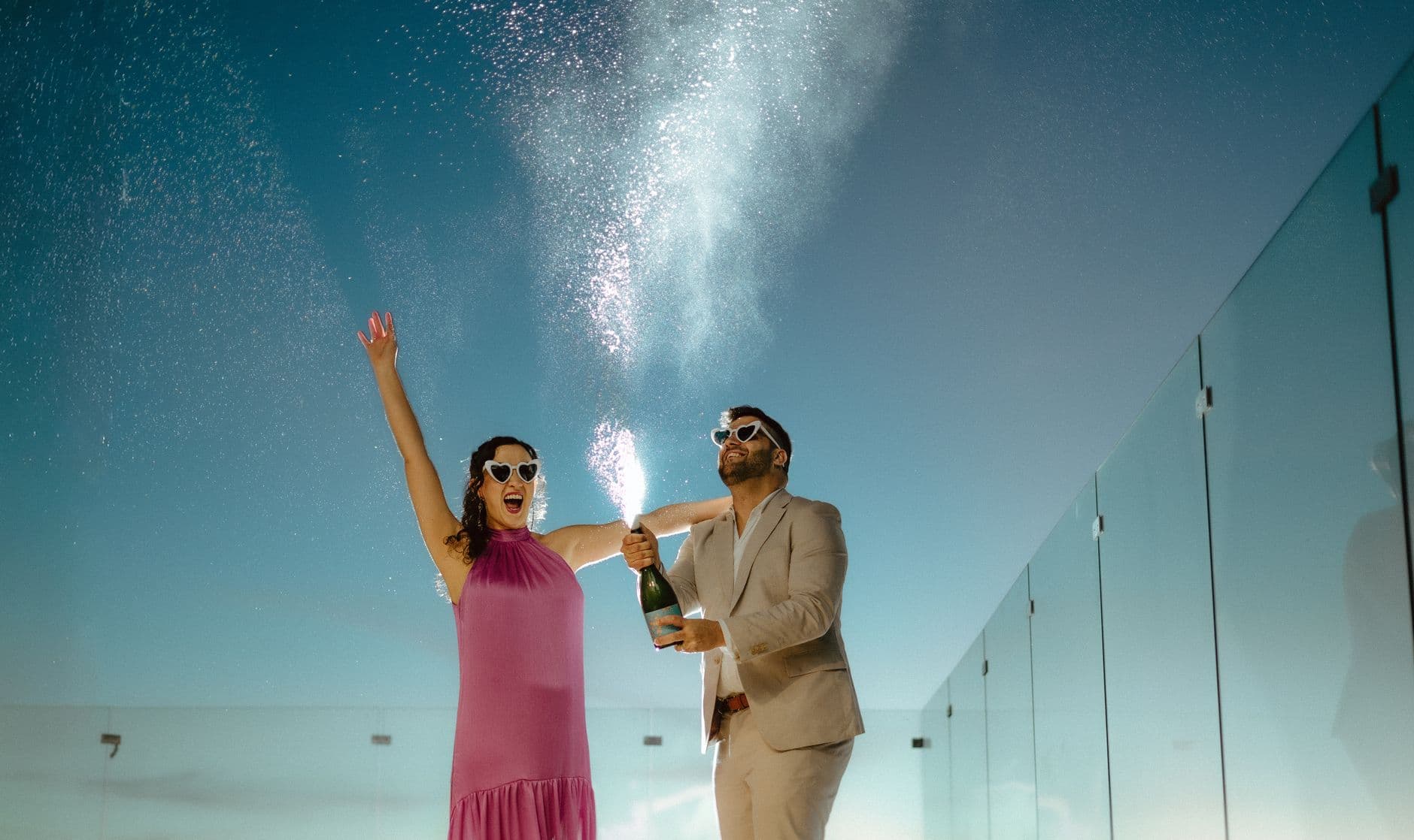 A woman in a pink dress cheers while a man in a beige suit sprays sparkling wine on a rooftop.