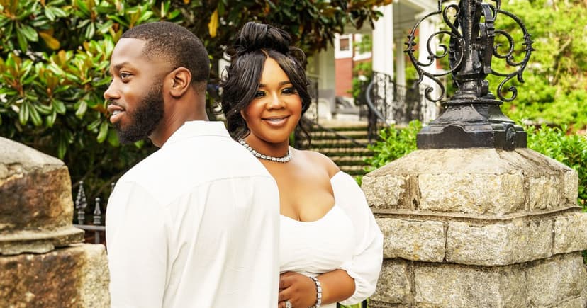 A well-dressed couple in white stand back to back beside a stone gatepost and iron lamp outside a grand old house.