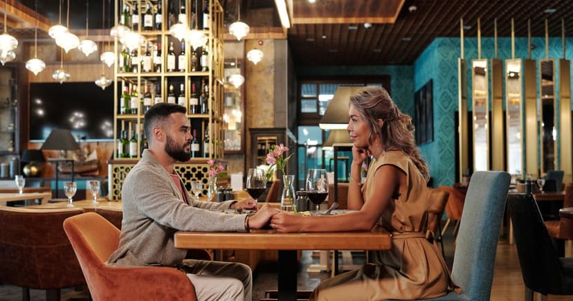 A man and a woman sit facing each other at a small restaurant table, hands touching beside two glasses of red wine.
