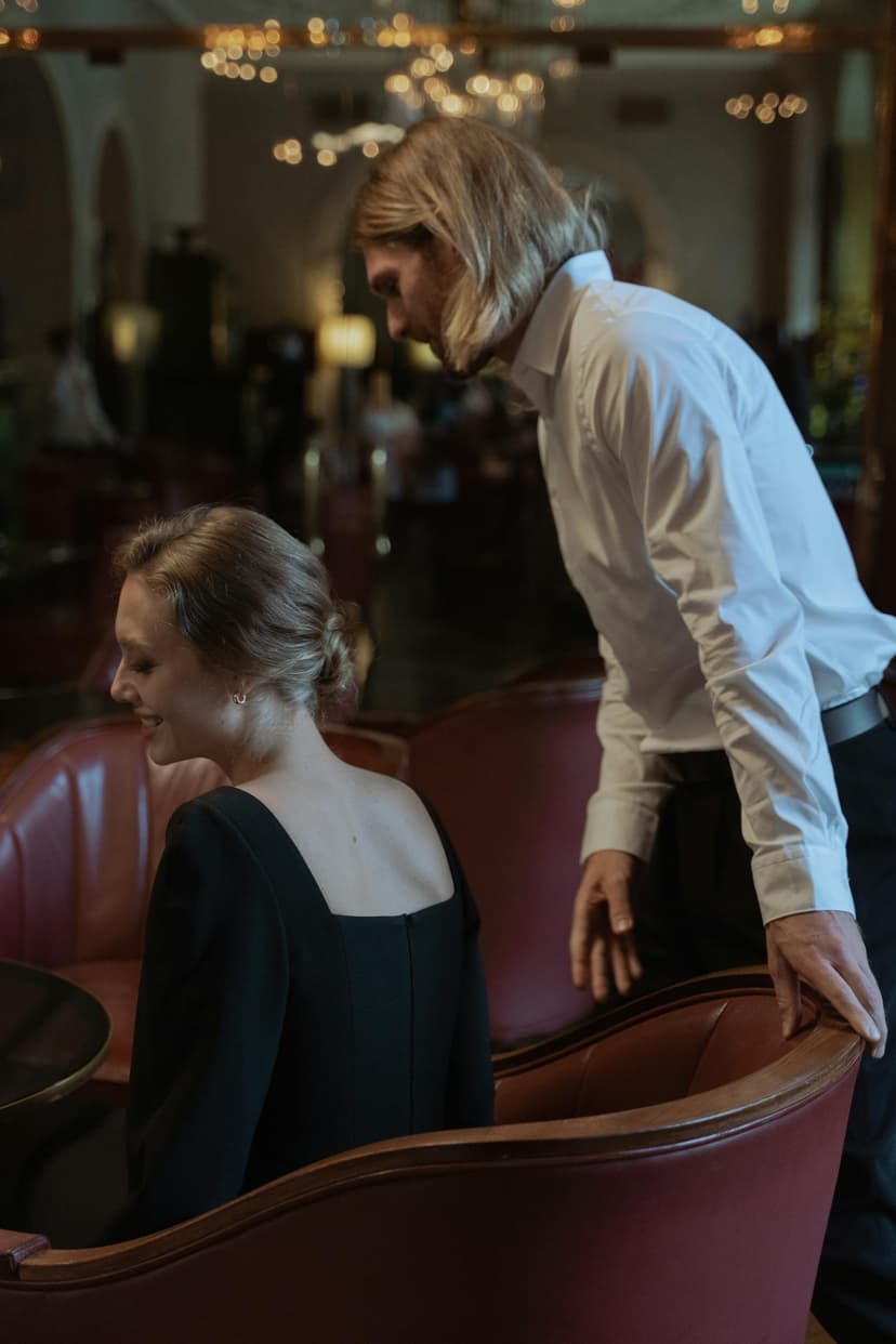 Frau im schwarzen Abendkleid an einem eleganten Restaurant-Tisch, Mann im weißen Hemd steht hinter ihr