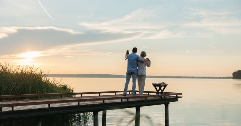 An older couple stand arm in arm at the end of a wooden dock, photographing a calm lake at sunset.