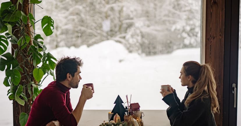 A man and a woman face each other with warm mugs at a window table, deep snow and snow-covered trees visible outside.