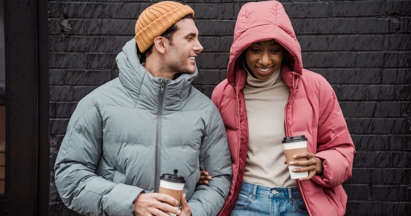 A man in a grey padded jacket and mustard knit hat and a woman in a rose-pink hooded parka stand against a black brick wall with takeaway coffee cups in their hands.