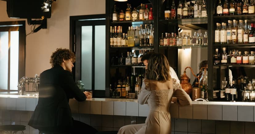 A man in a dark jacket and a woman in a long champagne satin dress lean on the same bar counter a seat apart, lit from behind by the shelves of bottles.