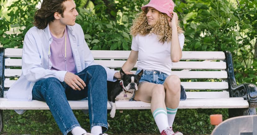 A man and a woman sit talking on a white park bench in summer greenery with a small dog between them.