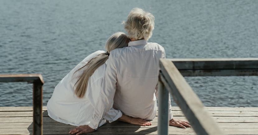 Two people in white shirts sit close together on a sunlit wooden dock, looking out over open water.