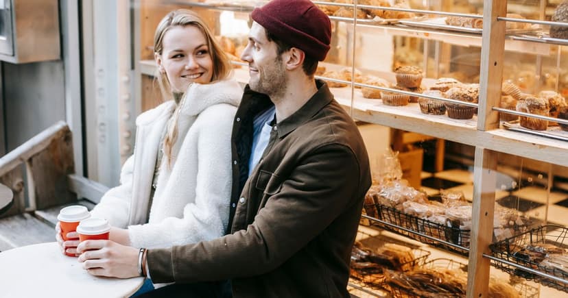 A woman in a cream fleece coat and a man in a brown chore jacket and maroon knit cap sitting with takeaway coffee at a bakery window counter.