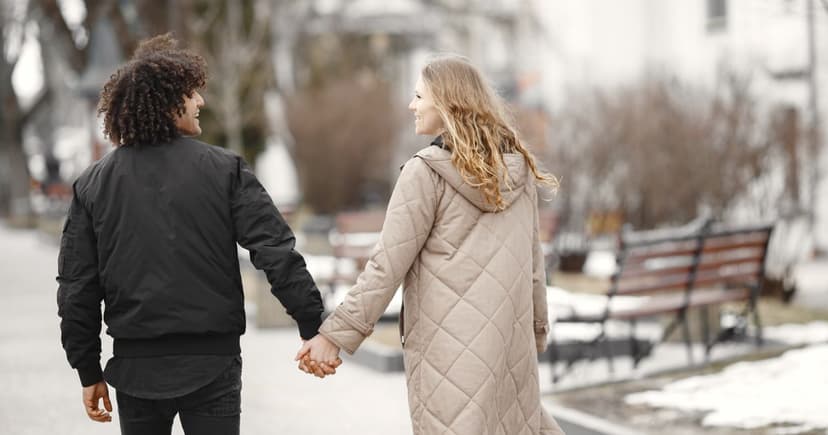A woman in a long beige quilted coat and a man in a black jacket holding hands on a snow-edged town path in flat winter light.