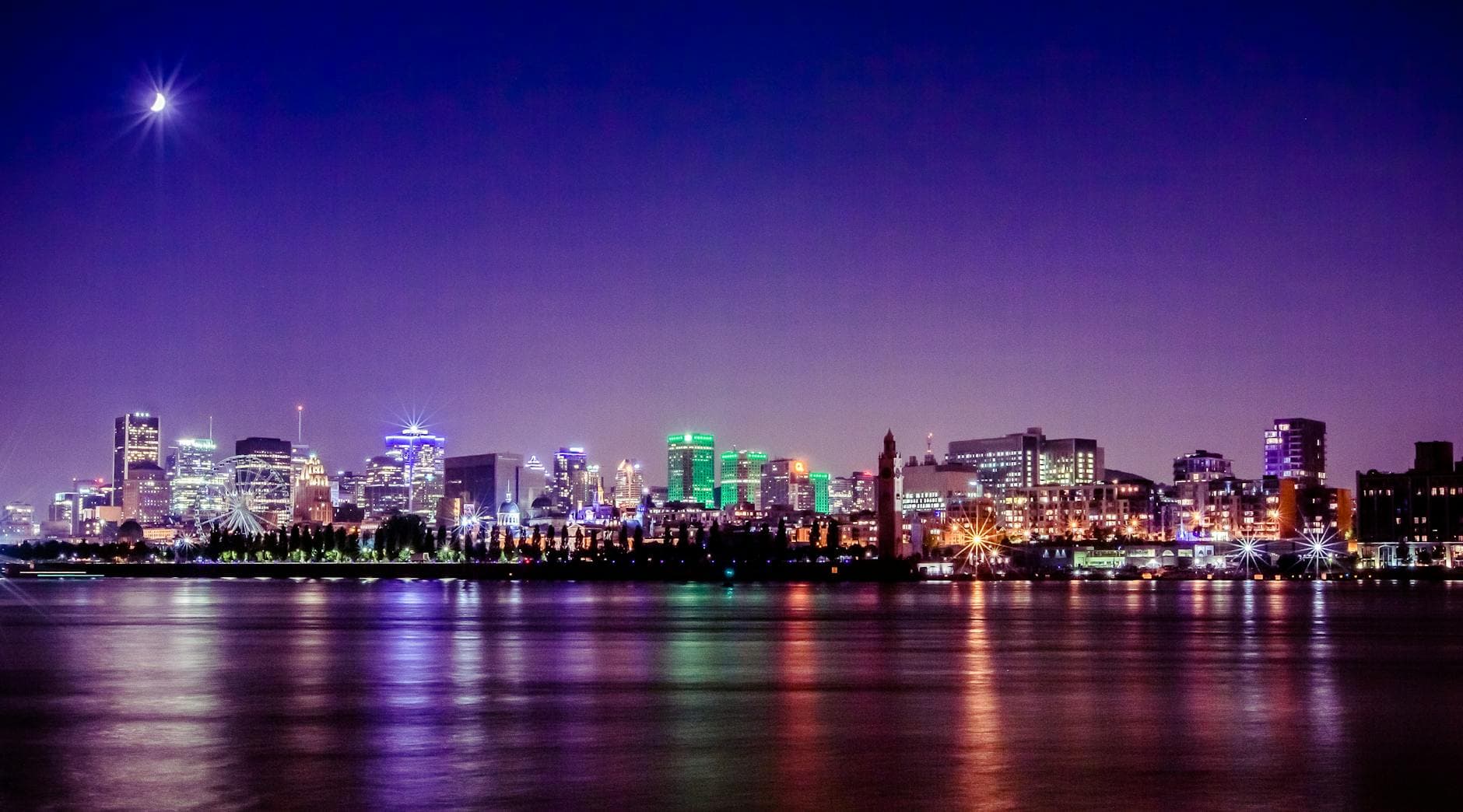 A lit city skyline and a crescent moon reflected in calm water at night.