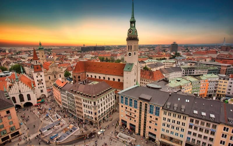Blick über Altstadtdächer und einen hohen Kirchturm bei orangefarbenem Sonnenuntergang.