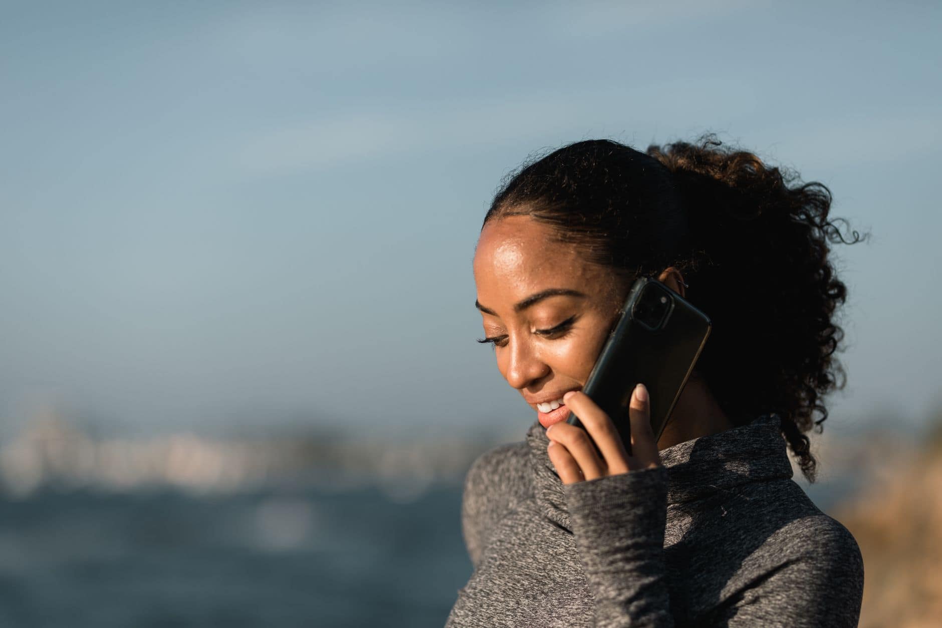 Uśmiechnięta kobieta z upiętymi lokami rozmawia przez telefon na tle błękitnej wody