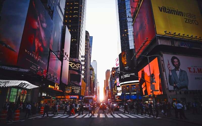Crowds cross a wide avenue between tall billboard-covered buildings at sunset.