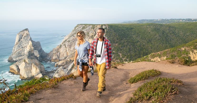 A couple walk together along a grassy clifftop path high above the ocean, with sea stacks in the water below.