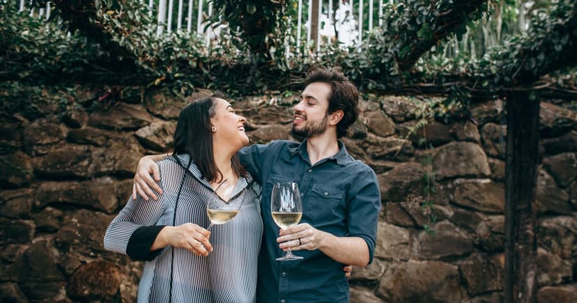 A couple laugh together holding glasses of white wine in front of a rough stone wall covered in greenery.