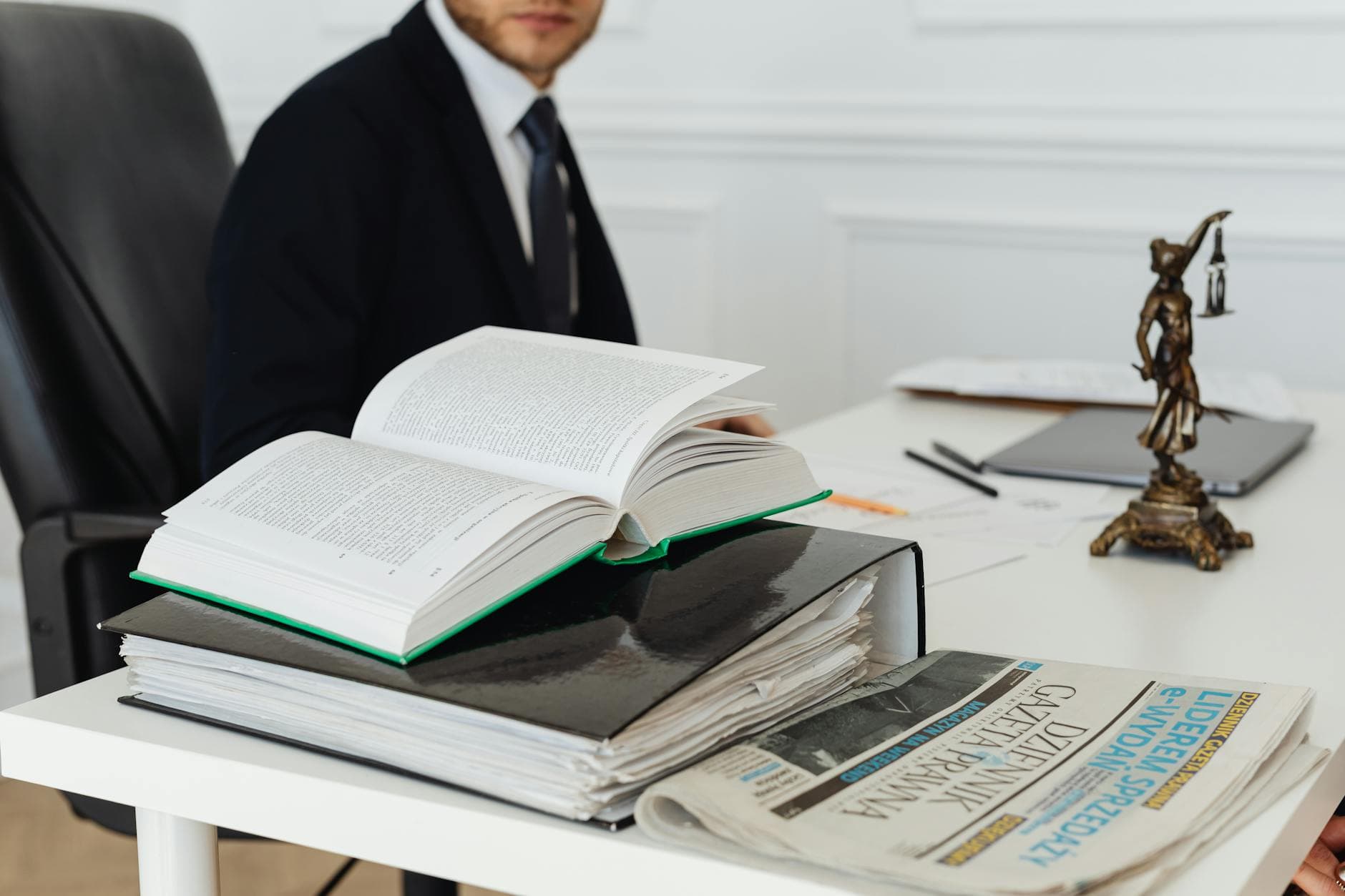 Mann i mørk dress sitter ved et skrivebord med oppslått bok, avis og en justitiafigur.