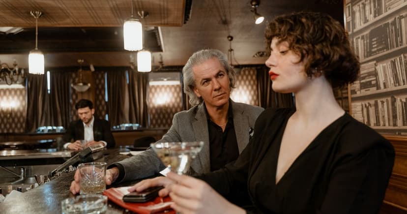 A silver-haired man in a grey jacket sits at a wooden bar counter beside a woman in black holding a martini glass.