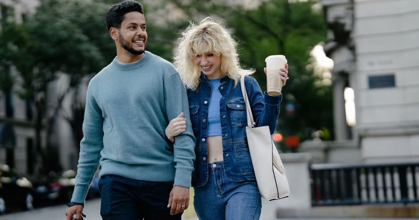 A smiling couple walk arm in arm past leafy stone buildings in daylight, the woman carrying a takeaway coffee and a tote bag.