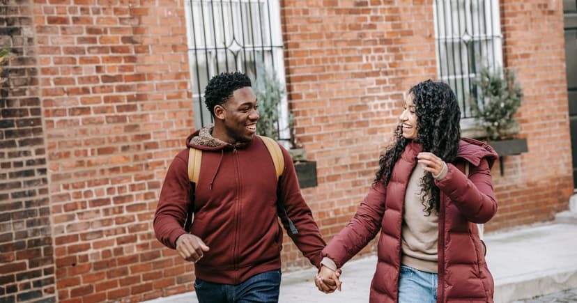 A man in a dark red hooded sweatshirt and a woman in a long quilted coat walk hand in hand along a sidewalk past red brick buildings, laughing at each other.