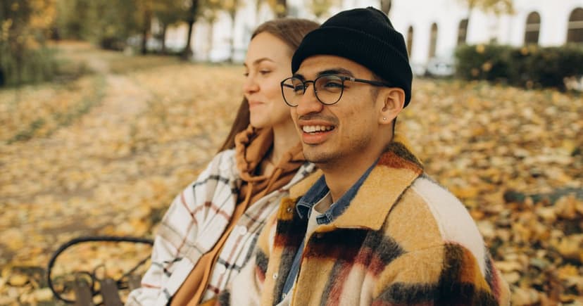 A man in a black beanie and a patterned wool jacket and a woman in a checked shirt and scarf sit side by side on a bench in a park covered in fallen leaves, both smiling at something off camera.