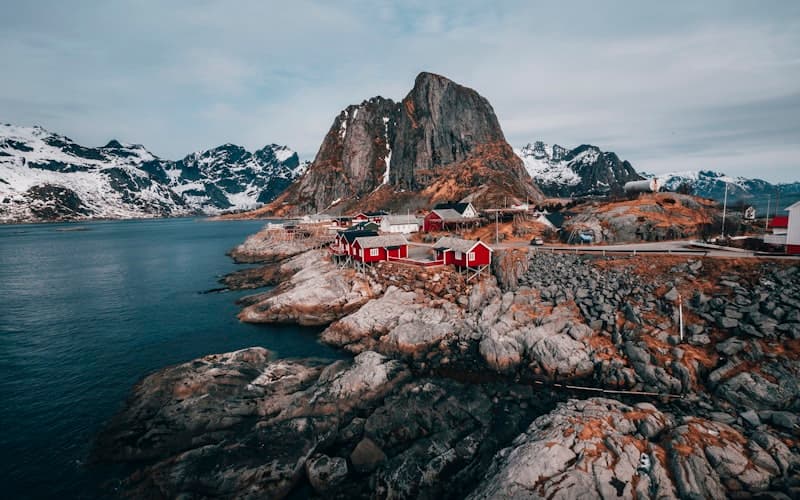 Røde trehus på en steinete odde med snødekte fjell og blågrønn fjord bak.