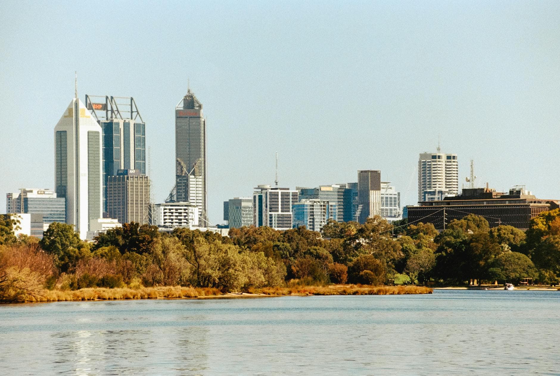 Office towers rise behind golden riverside trees across a wide, calm stretch of water.