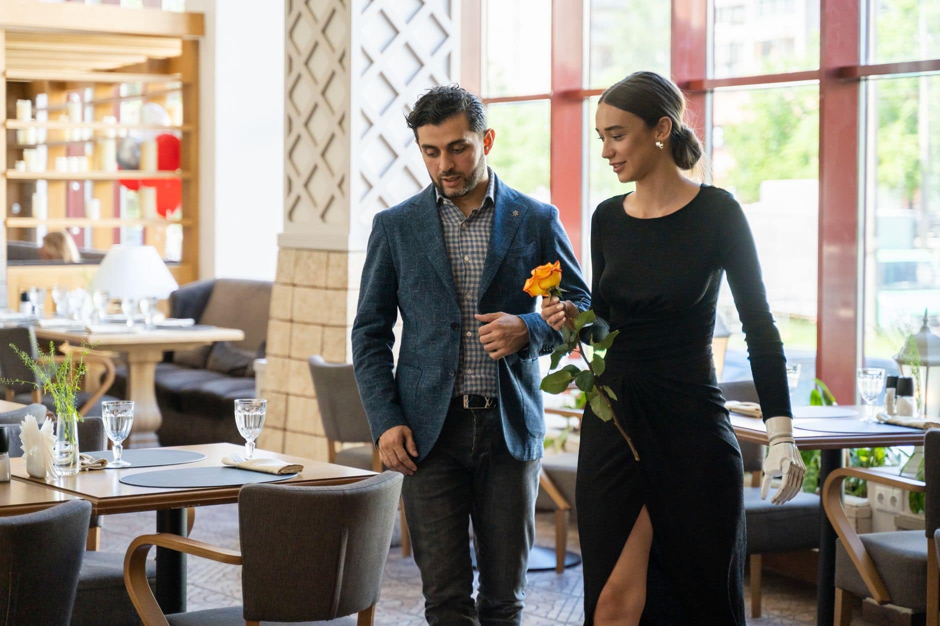 Elegancko ubrana para idzie przez jasną restaurację; kobieta trzyma pomarańczową różę