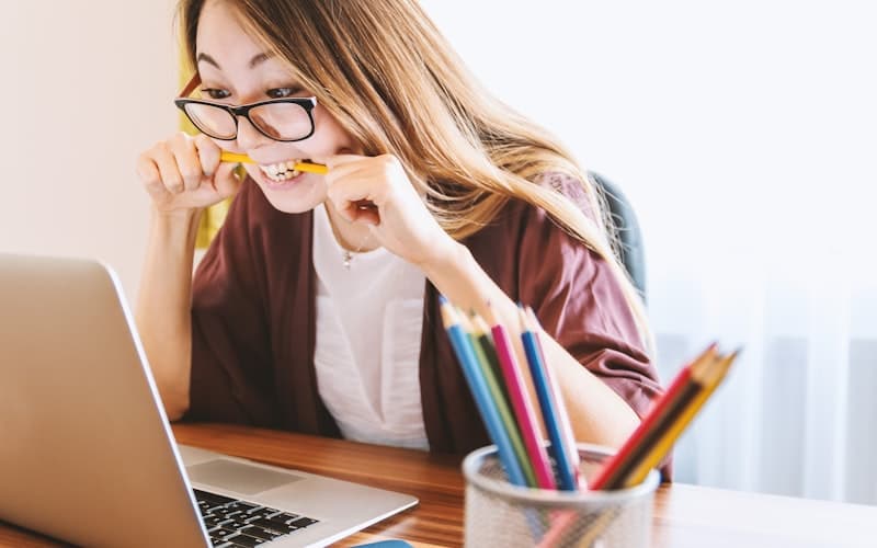 Vrouw met bril bijt op een geel potlood achter een laptop met kleurpotloden ernaast.