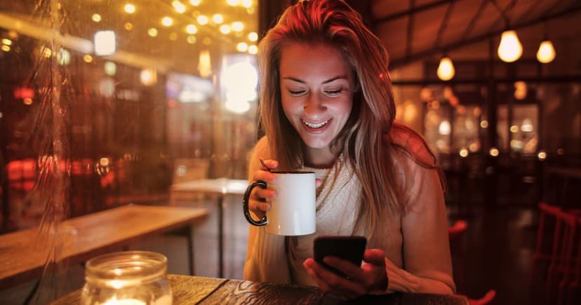 Smiling woman in a cream sweater reading her phone over a mug of coffee in a warmly lit cafe with string lights